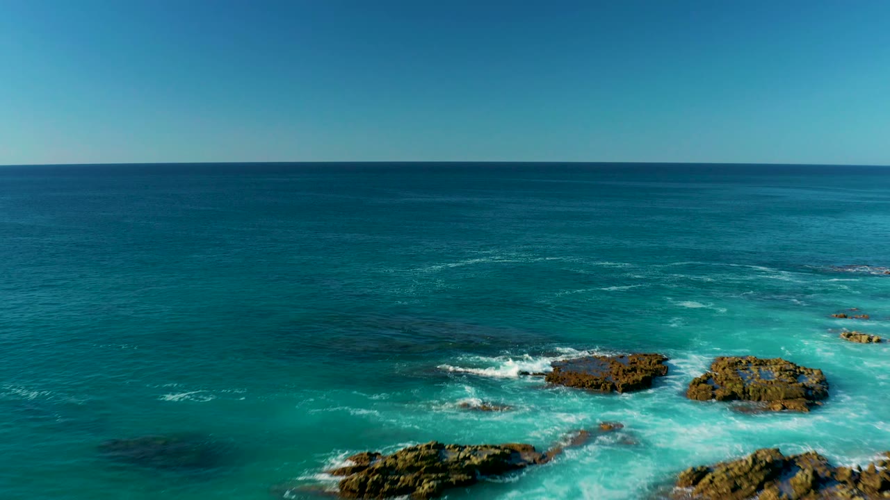 Aerial coastal view at La Fortuna, Baja California Sur, with rocks shifting toward the right side of the frame, white foam curling around them, and no visible people.
