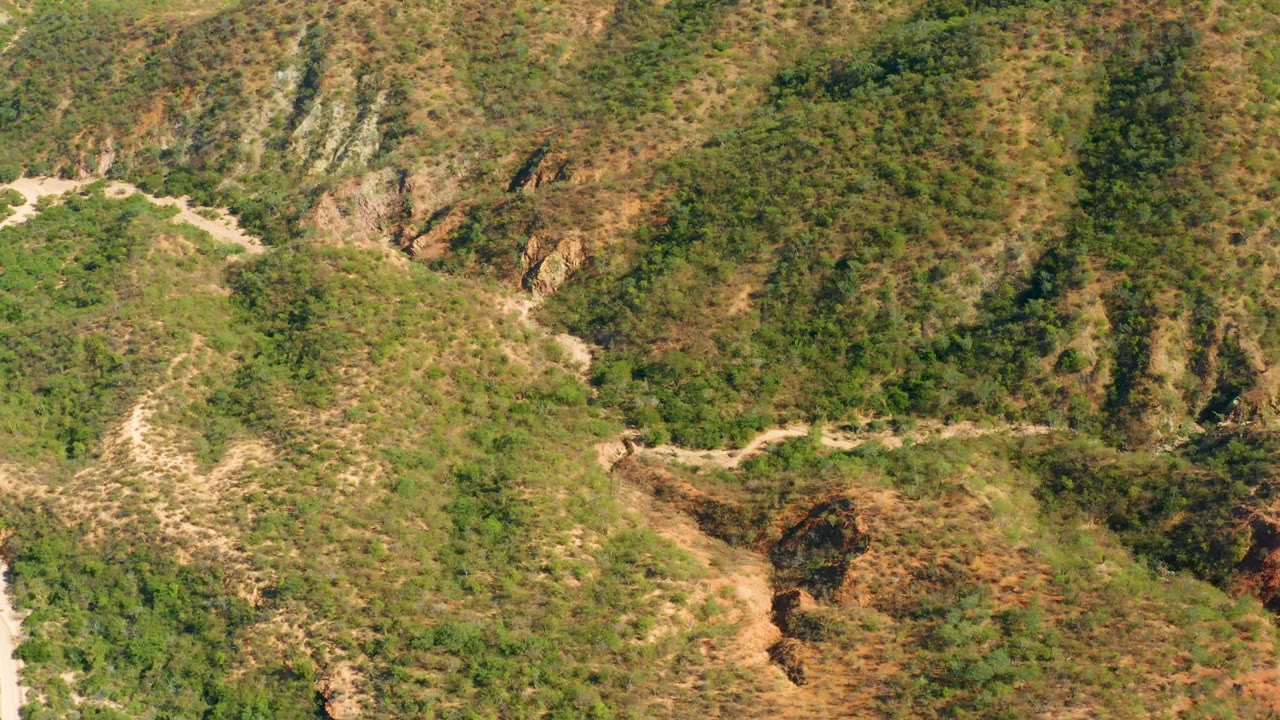 Aerial frame from El Salado Viejo Los Cabos showing a pale dirt road curving across scrub-covered hillsides with rocky ravines and no visible people.