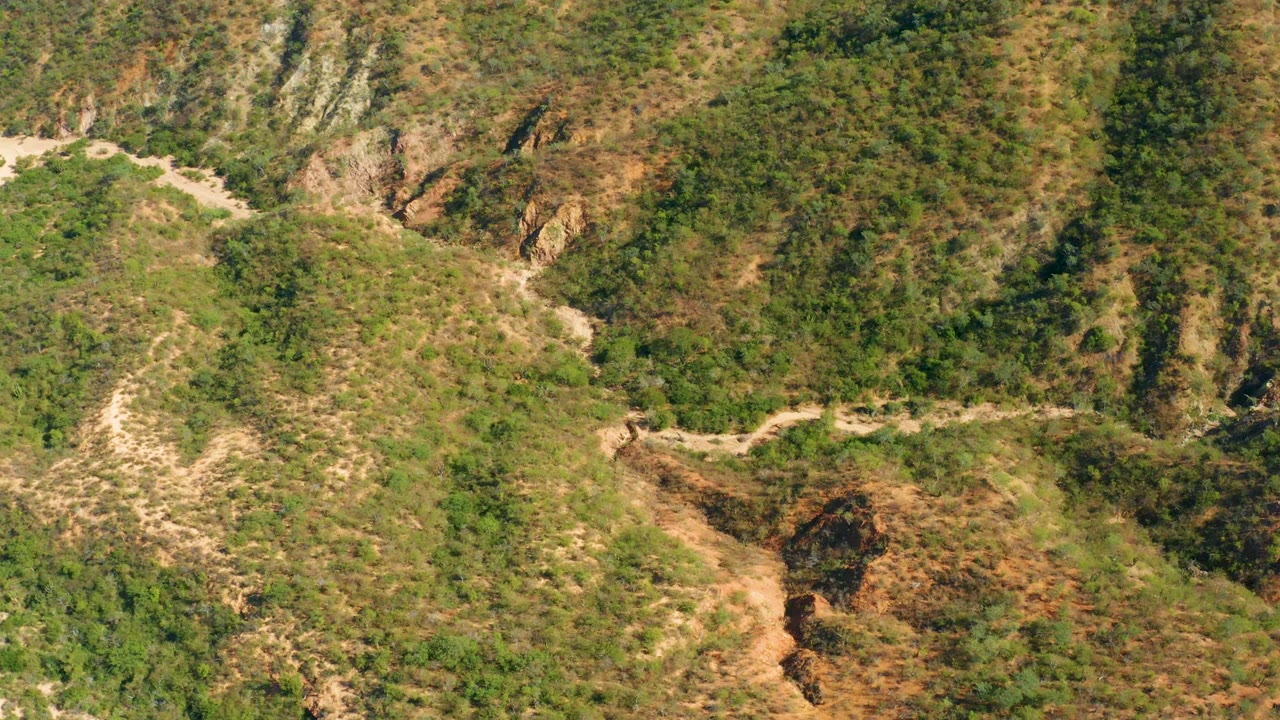 Elevated frame over El Salado Viejo Los Cabos showing the road bending near a rocky outcrop, with green brush and dry patches covering the slopes and no visible people.