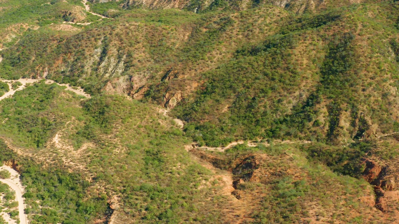 Aerial frame from El Salado Viejo, Los Cabos showing a pale road entering from the lower right below rocky ridges and mottled green vegetation, with no visible people.