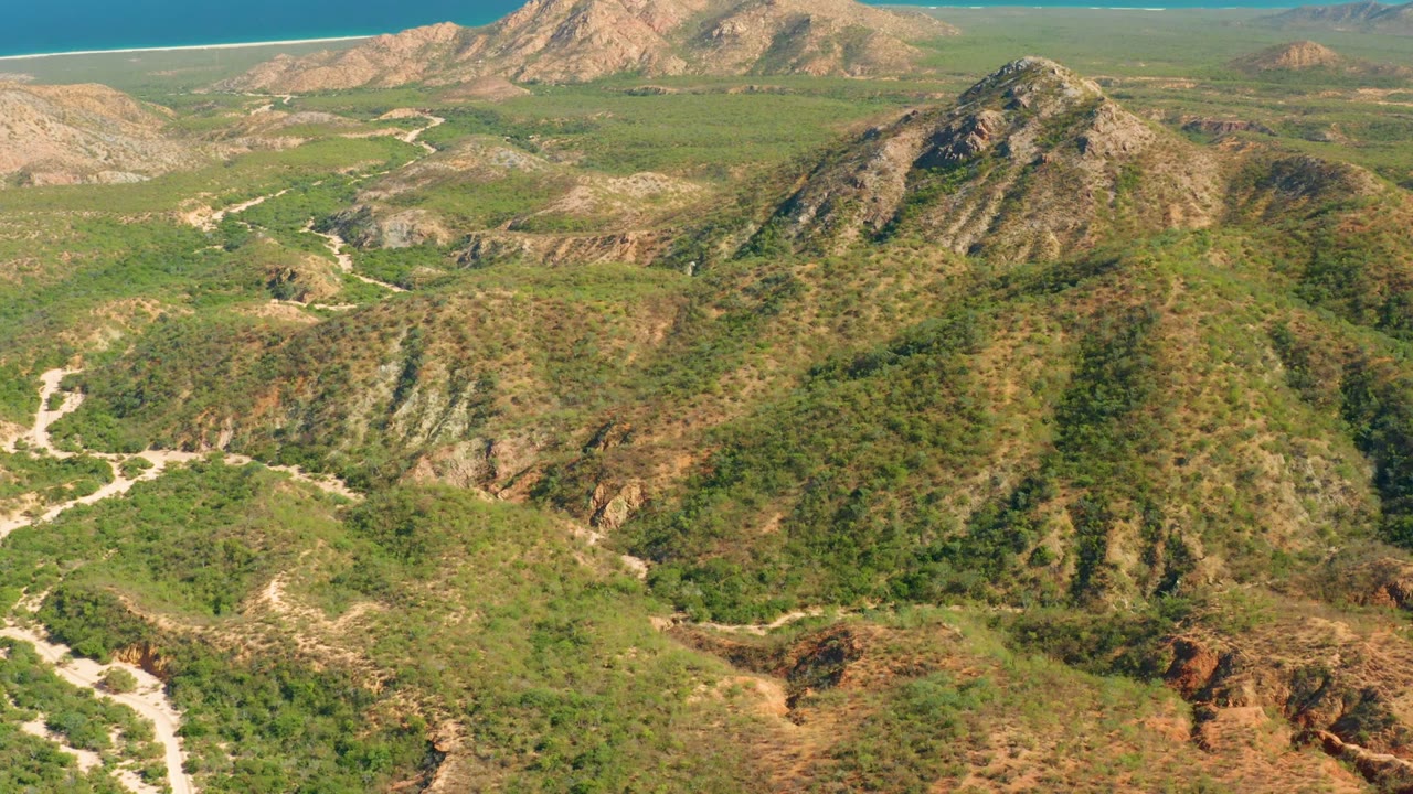 Aerial frame over El Salado Viejo Los Cabos, Baja California Sur, showing green hills, pale dirt paths, rocky ridges, reddish soil, distant ocean, and no visible people.