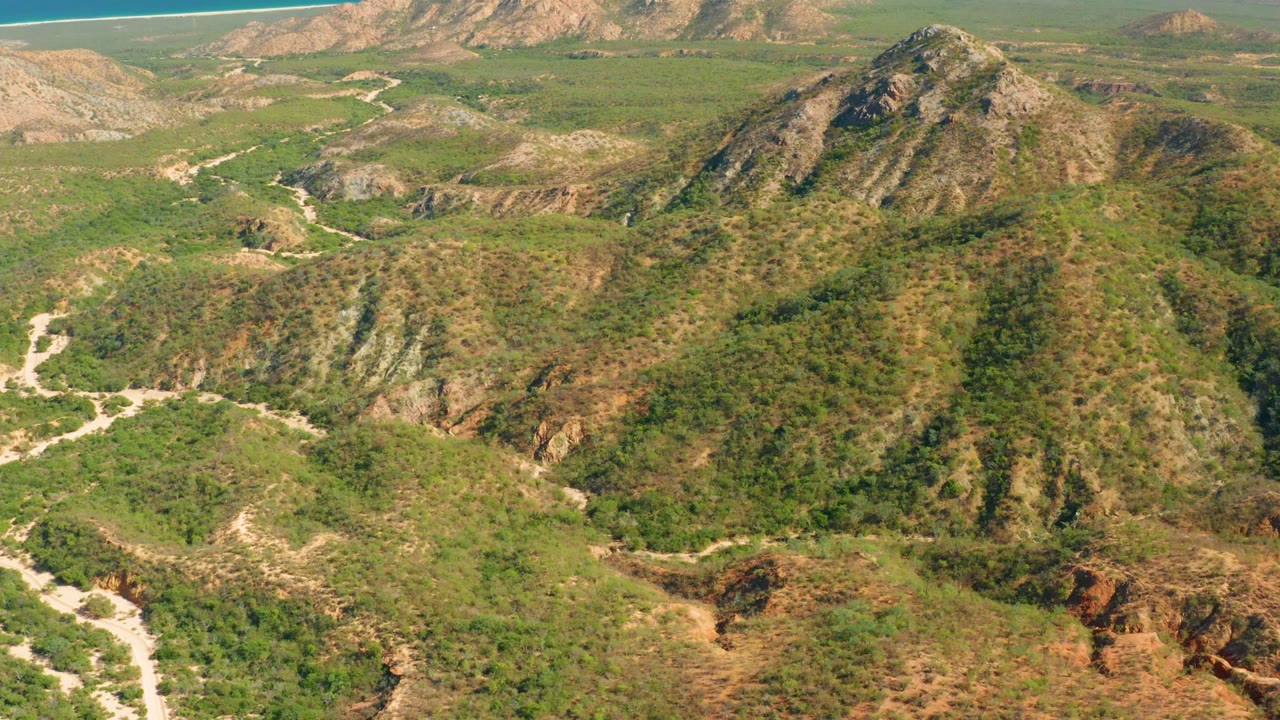 Aerial view over scrub-covered hills with a pale winding path and distant ocean strip.