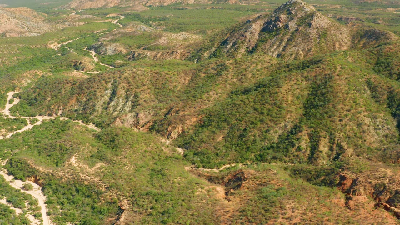 Drone-like frame over El Salado Viejo Los Cabos showing rugged green slopes, a pale road threading through valleys, reddish cuts in the earth, distant ocean, and no visible people.