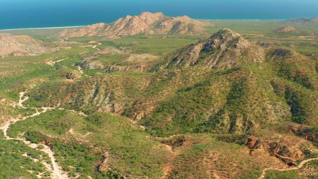 At 0:00, an aerial view over El Salado Viejo shows scrub-covered ridges in front, pale trails through the valleys, distant hills, a sandy shoreline, blue ocean, and no visible people.