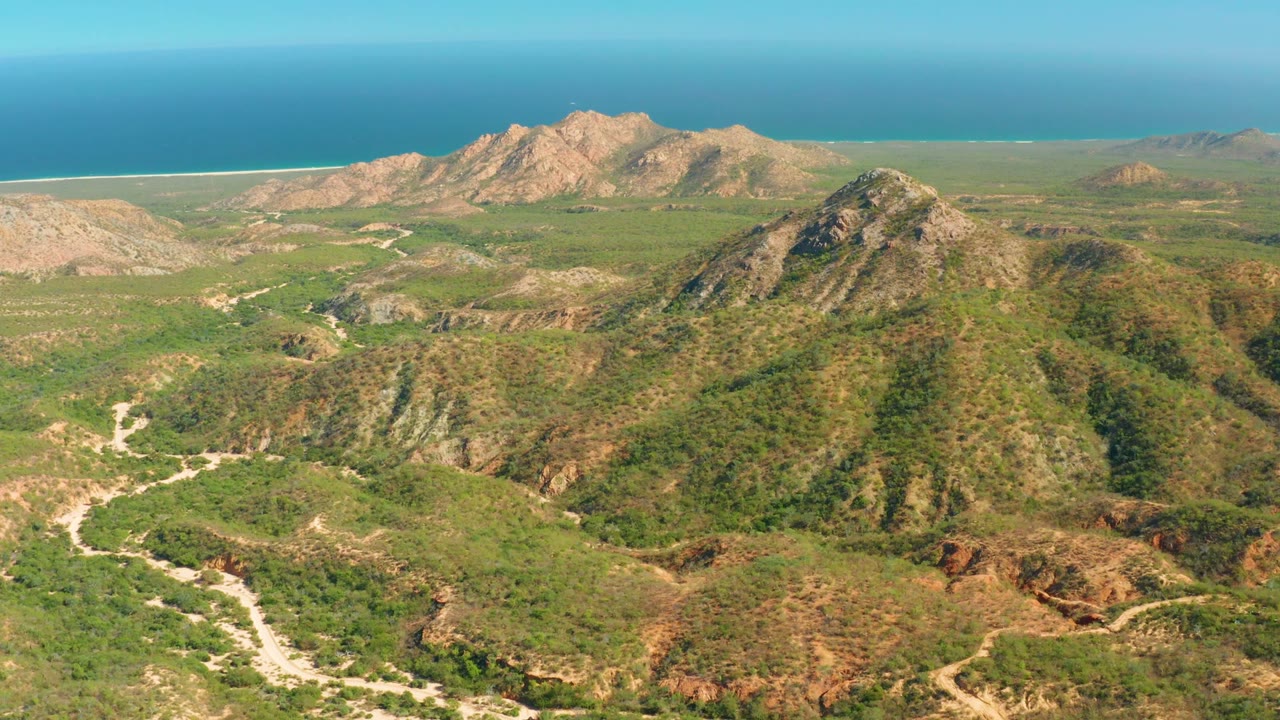A wide aerial frame over El Salado Viejo Los Cabos shows green hills, rocky mountain ridges, a pale winding dirt road, the ocean beyond the coastline, and no visible people.