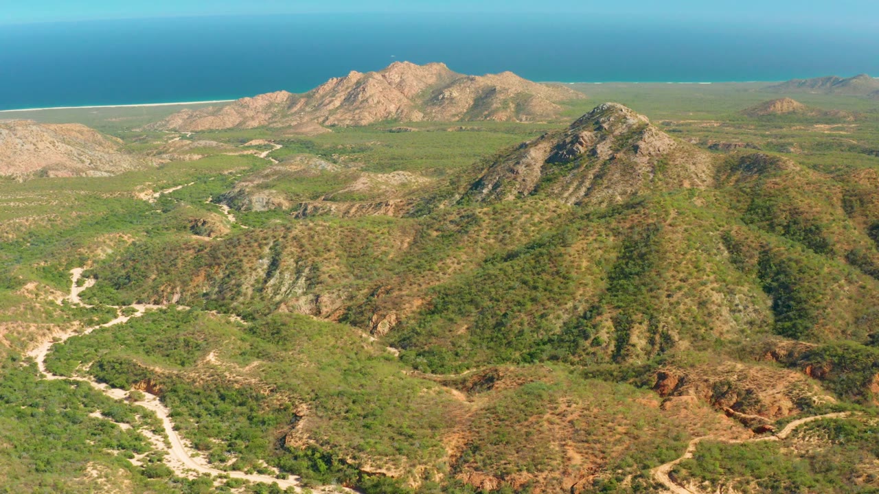 At 0:02, El Salado Viejo Los Cabos is seen from above with scrubby green slopes, a pale road bending through the terrain, distant rocky mountains, ocean water, and no visible people.