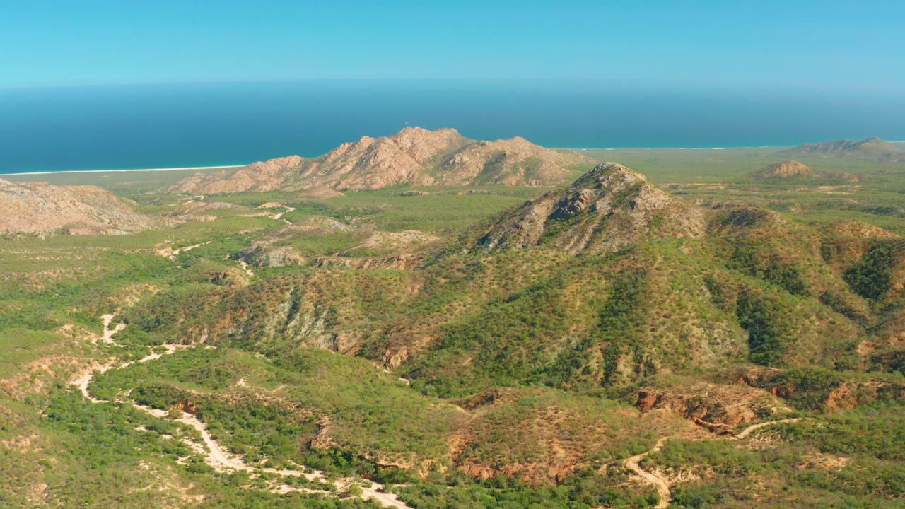Aerial frame from El Salado Viejo Los Cabos showing green hills with pale winding roads, rocky ridges beyond, a blue ocean horizon, and no visible people.