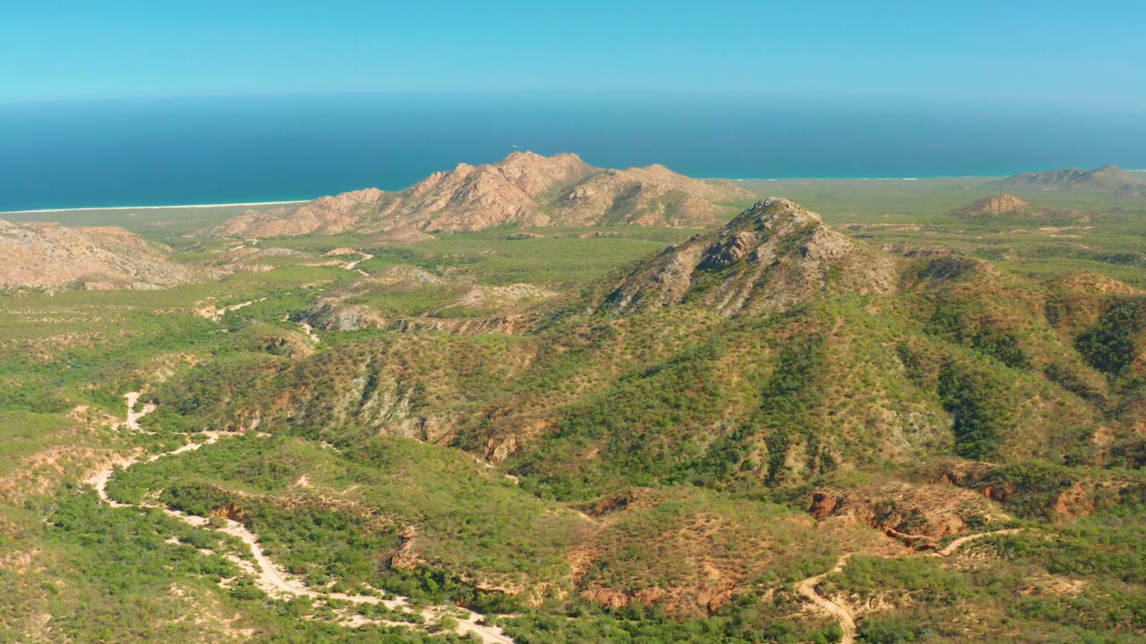 High aerial view of scrubby hills, pale dirt tracks, distant mountains, and blue ocean.