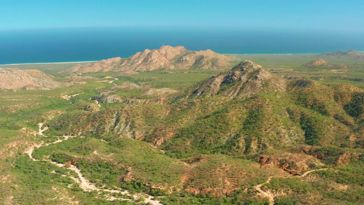 Aerial frame from El Salado Viejo Los Cabos centered on a rounded green ridge, with winding dirt roads below, rocky peaks behind, blue ocean, and no visible people.