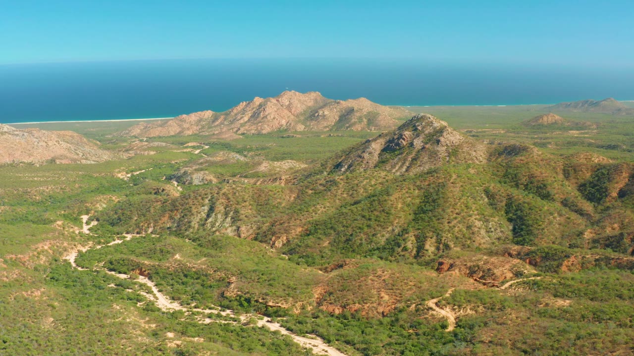 High aerial view across scrub-covered hills toward rocky mountains and ocean.