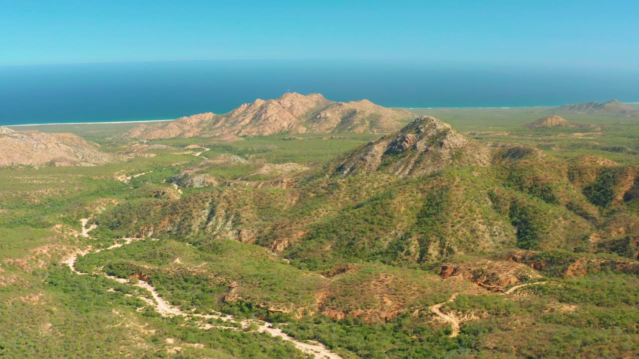 At 0:02 the drone perspective over El Salado Viejo shows the winding track more clearly in the lower center, with layered green hills, rocky ridgelines, distant coast, and no visible people.