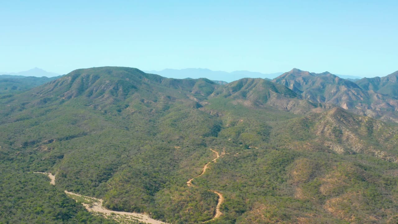 Aerial view over El Salado Viejo Los Cabos, Baja California Sur, with green hills, winding dirt roads, distant mountain ridges, clear sky, and no visible people.