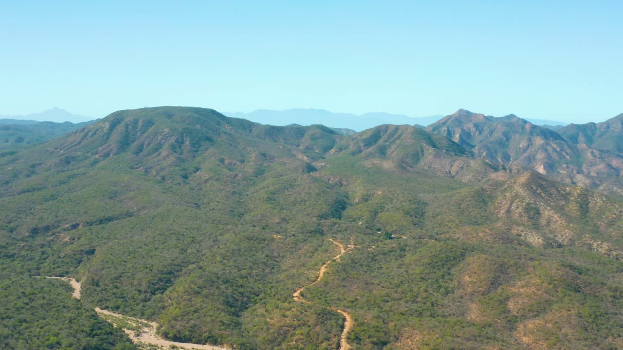 Aerial view in El Salado Viejo Los Cabos, Baja California Sur, with the winding dirt road lower in frame, green valleys in the midground, distant ridges, and no visible people.