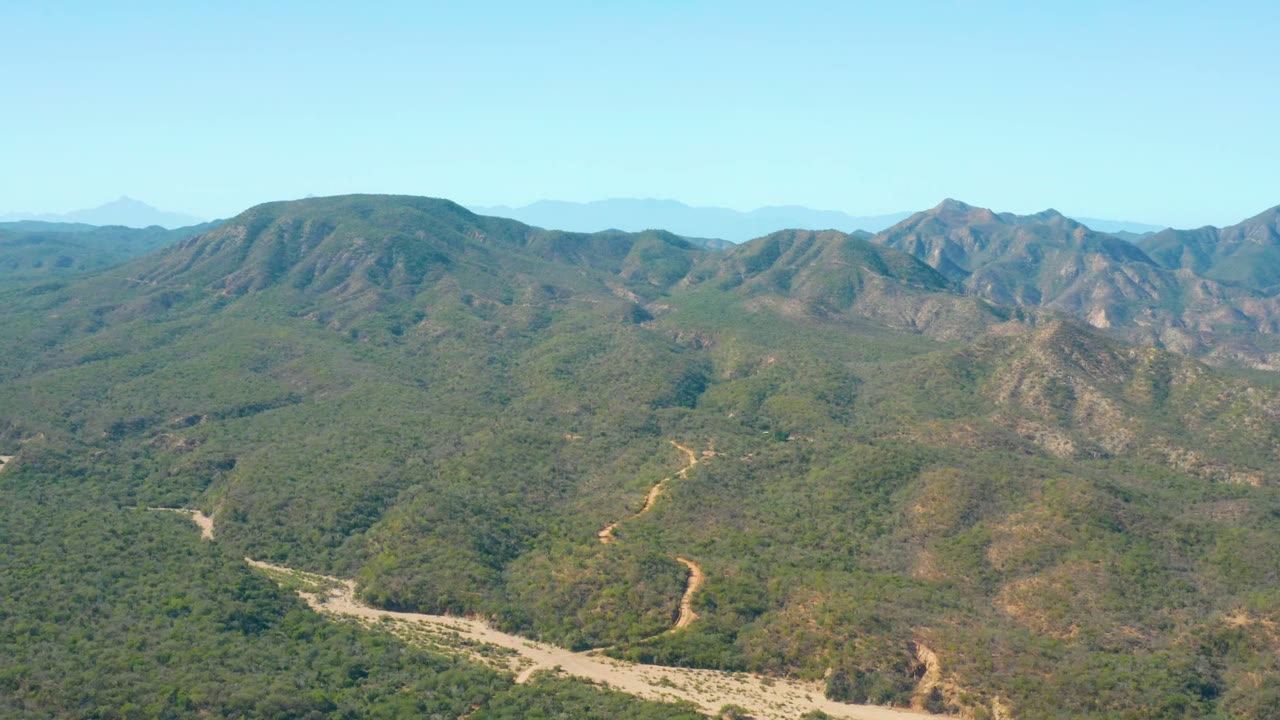 Wide aerial view over El Salado Viejo Los Cabos, Mexico, showing green hills, a pale winding riverbed at the lower left, distant ridges, clear blue sky, and no visible people.