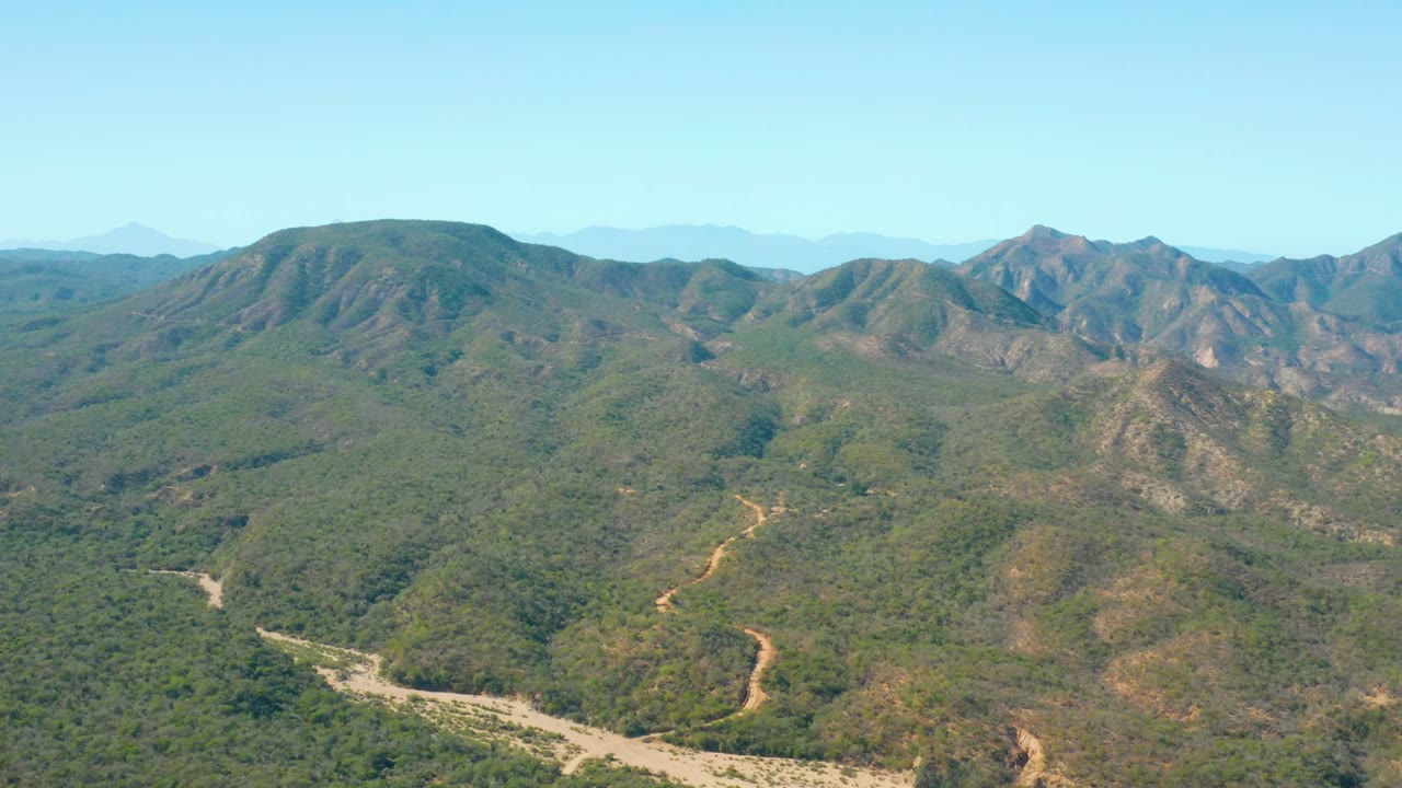 Wide aerial view in El Salado Viejo Los Cabos, Mexico, showing a pale winding channel along the valley floor, green-brown slopes, distant blue ridges, open sky, and no visible people.