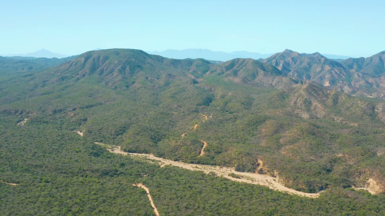 At 0:00, an aerial view over El Salado Viejo Los Cabos shows a pale winding river channel crossing green hills toward distant rugged mountains under a clear blue sky, with no visible people.