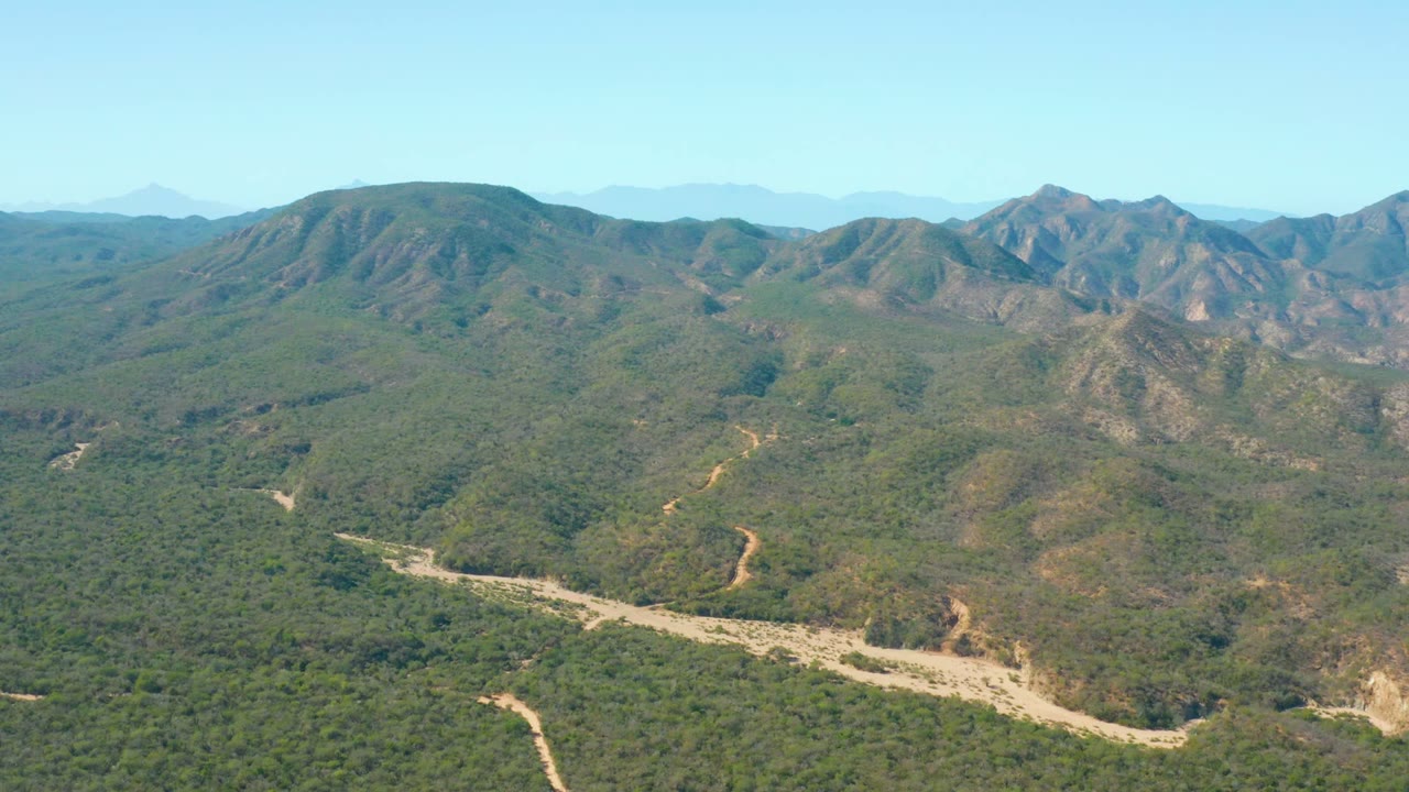 At 0:02, the aerial composition shows El Salado Viejo Los Cabos mountain terrain with a light-colored winding riverbed cutting across the lower midground and rocky ridges rising in the background, with no visible people.