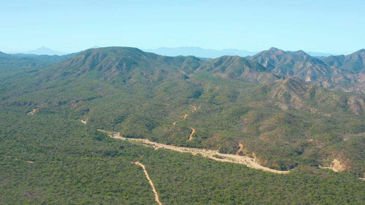 Wide aerial frame over El Salado Viejo Los Cabos shows a green valley, pale riverbed, winding dirt tracks, rocky hills, blue sky, and no visible people.