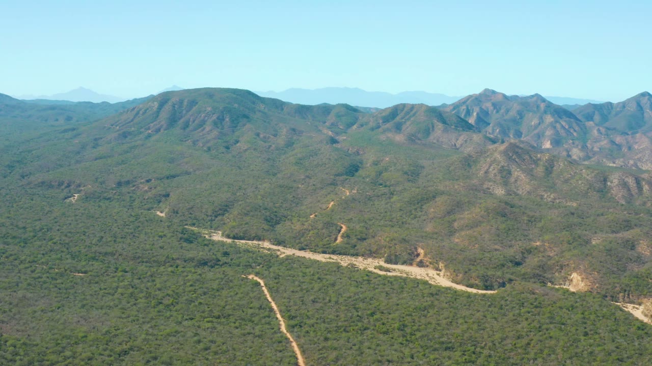 Opening frame of El Salado Viejo Los Cabos showing dense green terrain, pale dirt tracks, distant brown mountain slopes, clear blue sky, and no visible people.