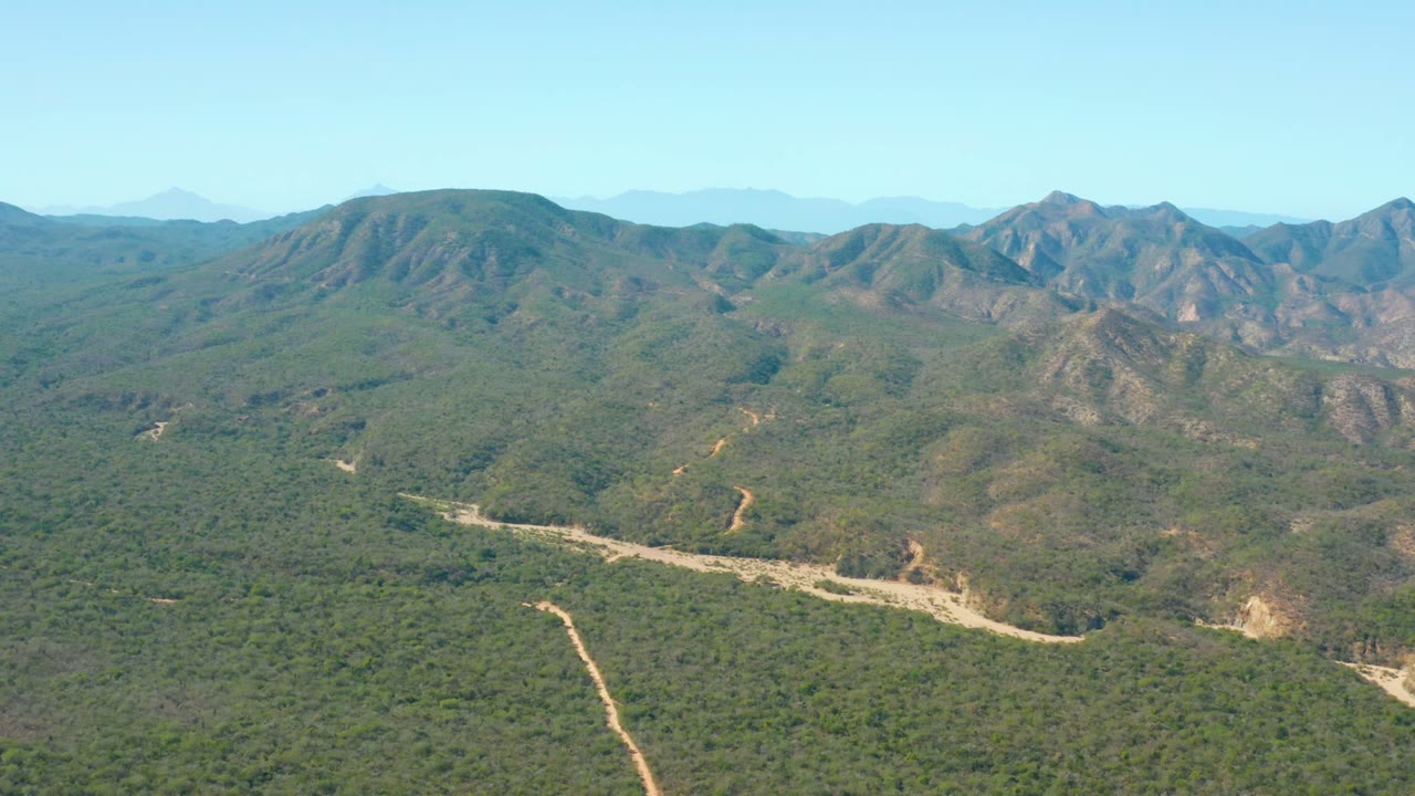 Static aerial landscape with forested hills and winding dirt tracks below mountains.