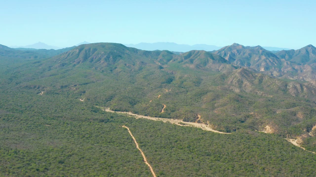 Aerial view over El Salado Viejo, Los Cabos, with a pale dirt road crossing green hills toward distant mountain ridges and no visible people.