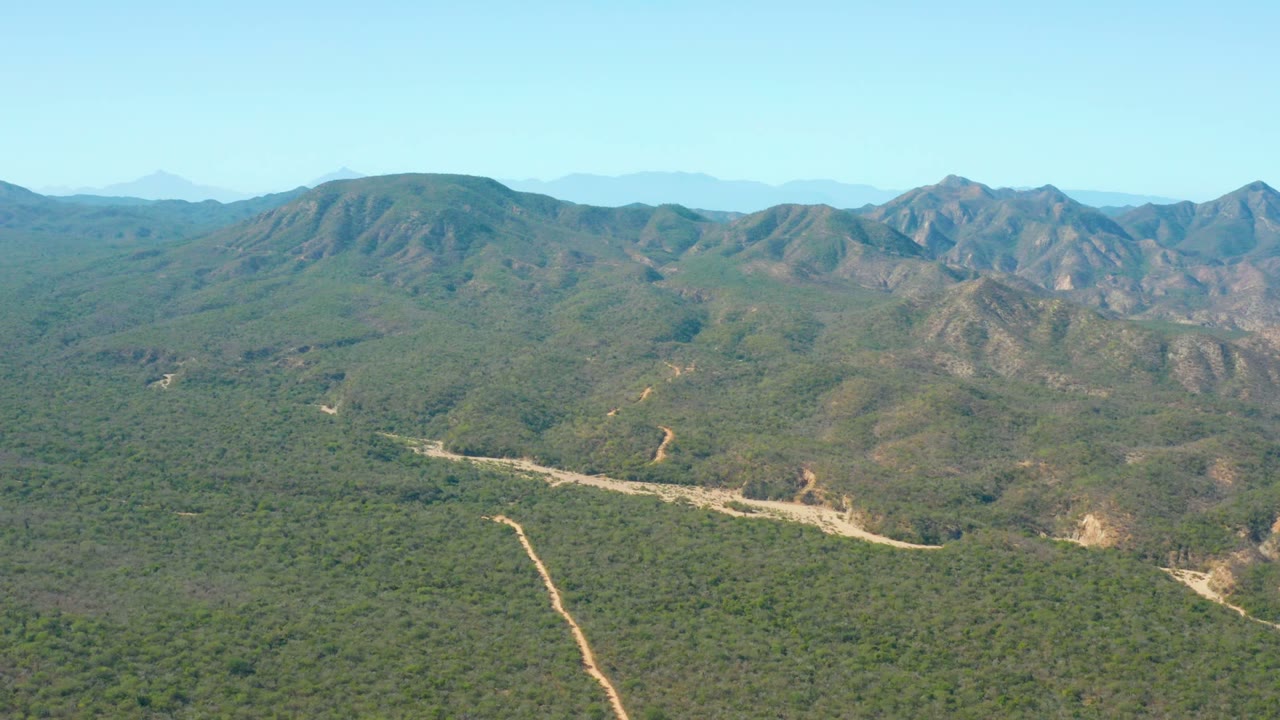 Wide aerial landscape with a dirt road cutting through dense green terrain and low mountains beyond.