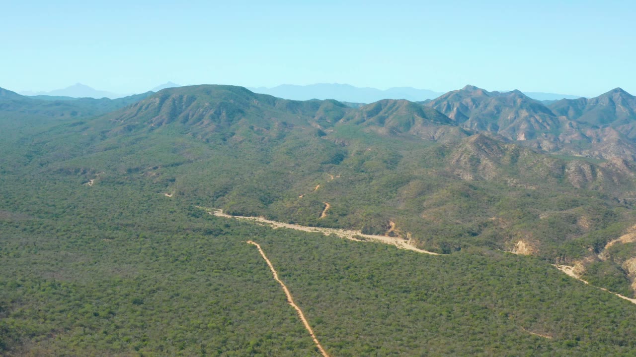 Wide aerial landscape with a pale road cutting through green hills toward rocky mountains.