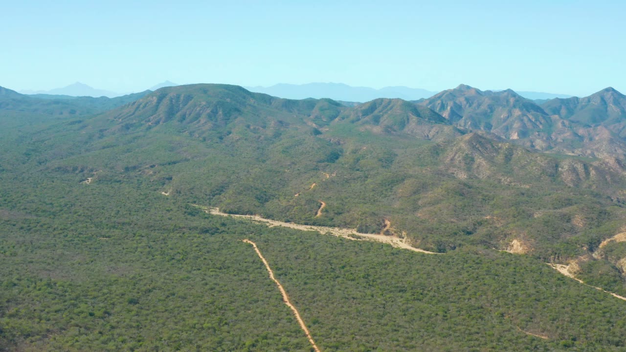 Aerial view over El Salado Viejo Los Cabos with a winding dirt road across scrub-covered hills, muted rocky mountains in the background, bright daylight, and no visible people.