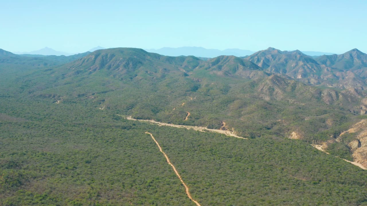 Aerial view over El Salado Viejo Los Cabos, Baja California Sur, with a pale dirt road cutting through green vegetation, mountain ridges on the horizon, and no visible people.
