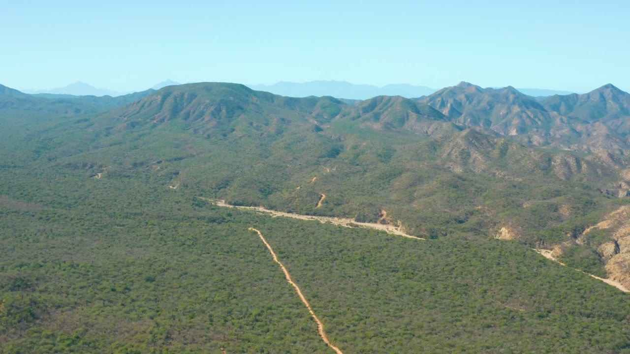 Wide aerial landscape with a dirt road crossing dense vegetation below mountains.