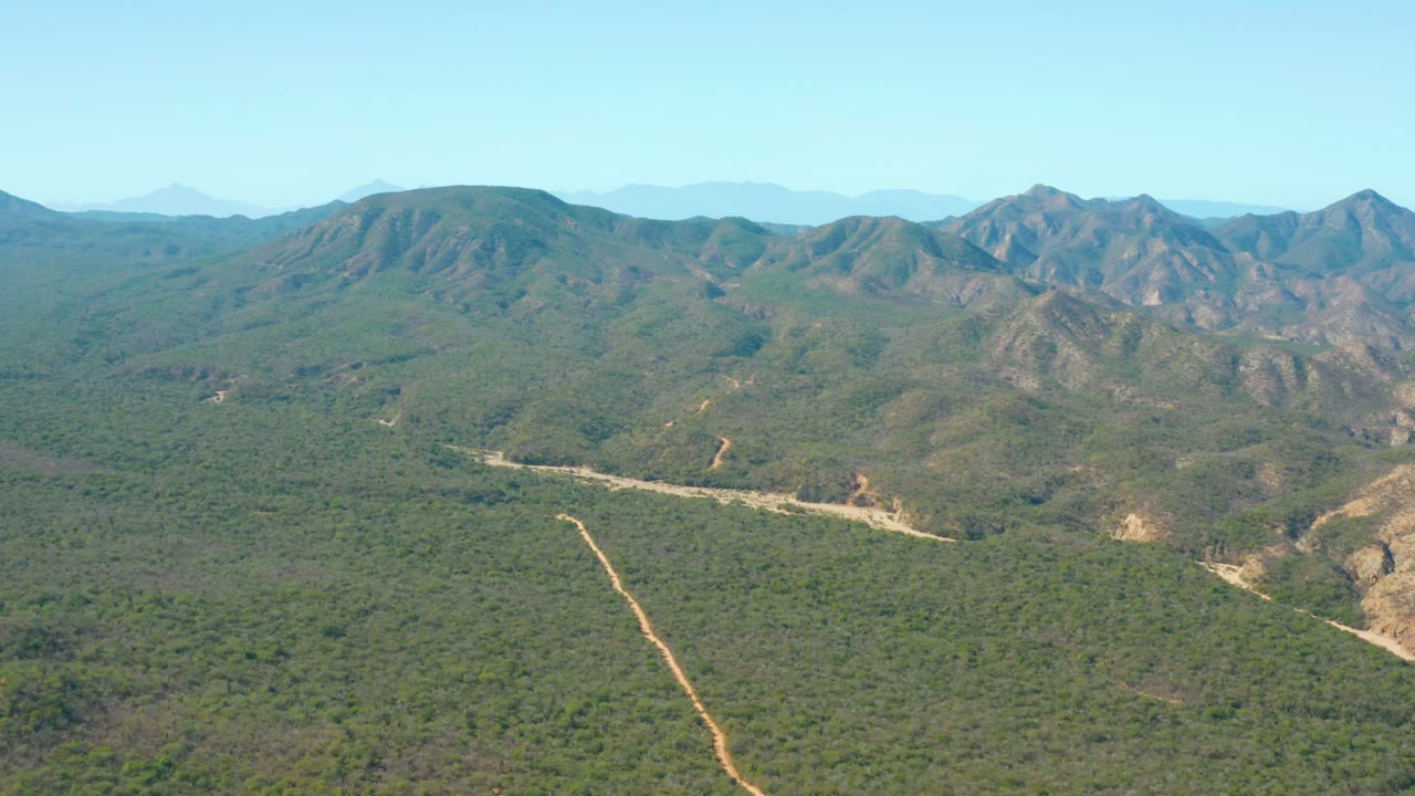 The frame shows El Salado Viejo Los Cabos terrain from above, with a pale dirt track in the lower valley, layered green hills, rocky mountain slopes, blue sky, and no visible people.
