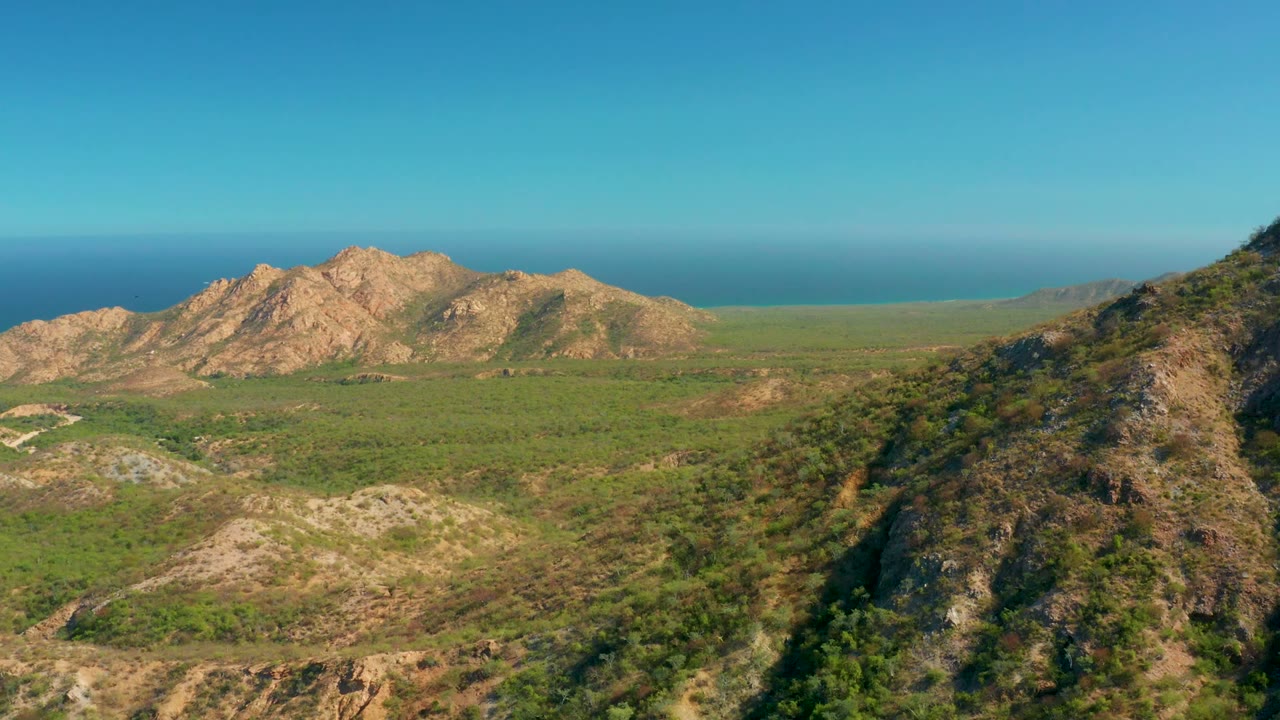 Opening aerial view over El Salado Viejo Los Cabos, with a dark foreground ridge, green valley floor, tan rocky hills, distant blue ocean, clear sky, and no visible people.