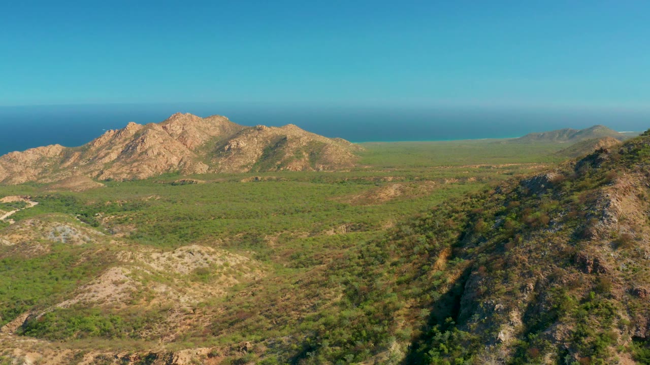 Elevated El Salado Viejo Los Cabos view with the foreground ridge filling the right side, green terrain across the middle, tan mountains to the left, distant ocean, and no visible people.
