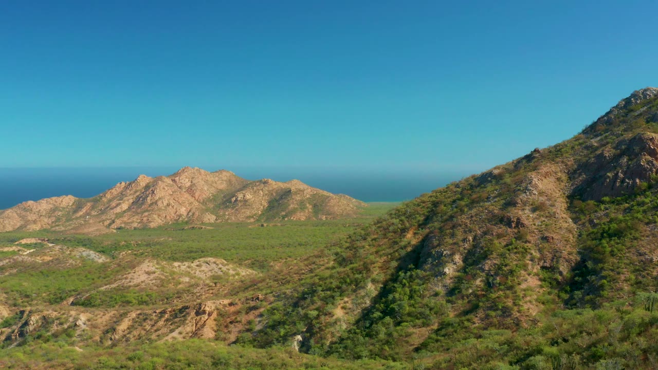 Wide aerial frame from El Salado Viejo Los Cabos showing rocky green hills in the foreground, tan mountain ridges across the valley, blue ocean on the horizon, clear sky, and no visible people.