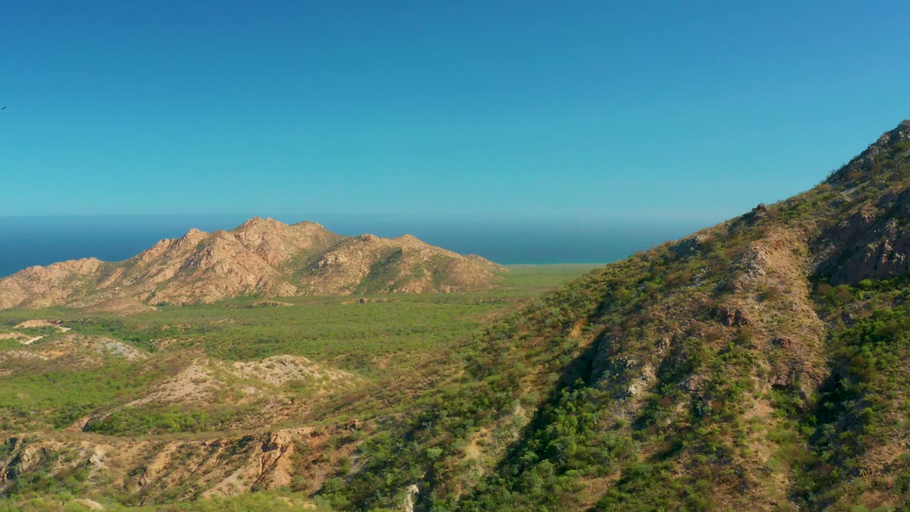 El Salado Viejo Los Cabos landscape frame with tan ridges centered across the valley, green scrubland below, the near hillside clipped at frame right, ocean behind the mountains, and no visible people.