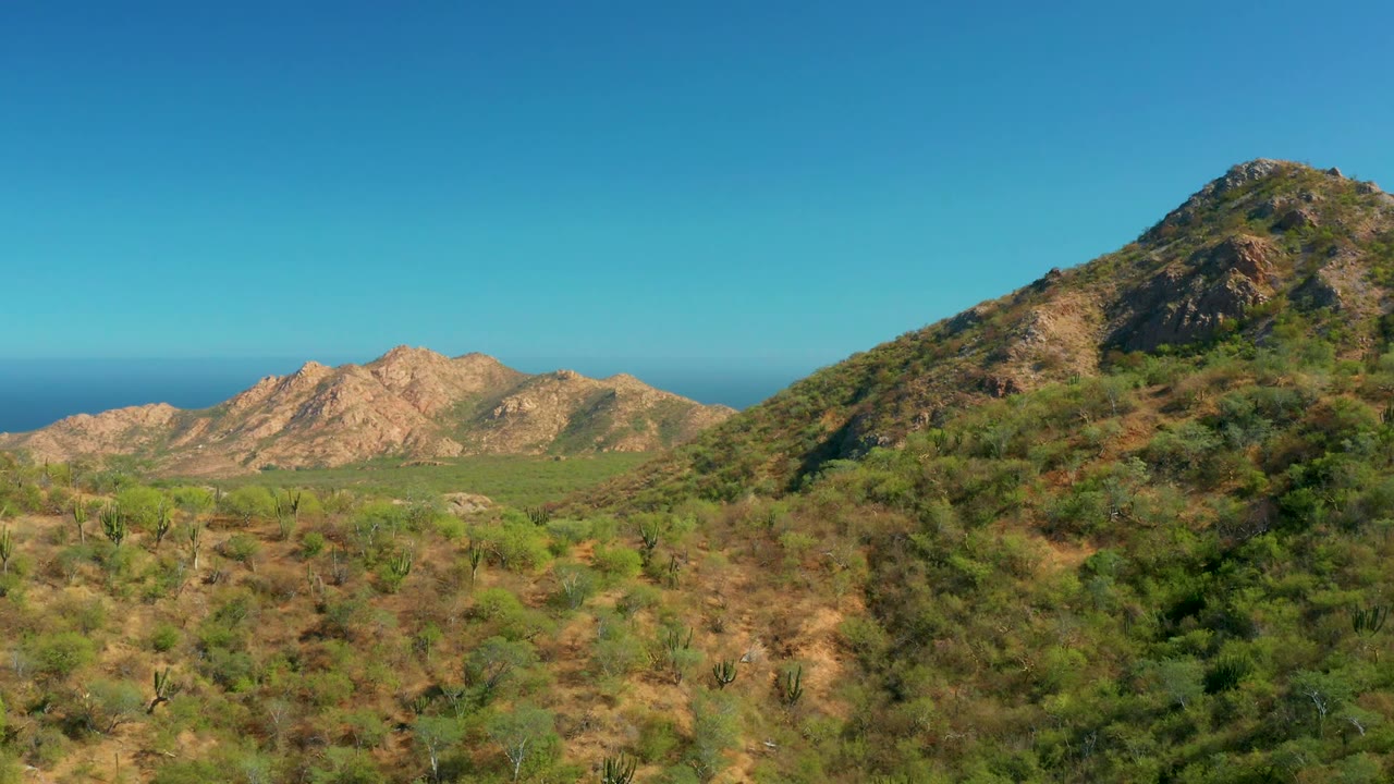 Opening view of El Salado Viejo Los Cabos with scrubby desert foreground, rocky hills across the valley, a larger slope at right, distant ocean, clear blue sky, and no visible people.
