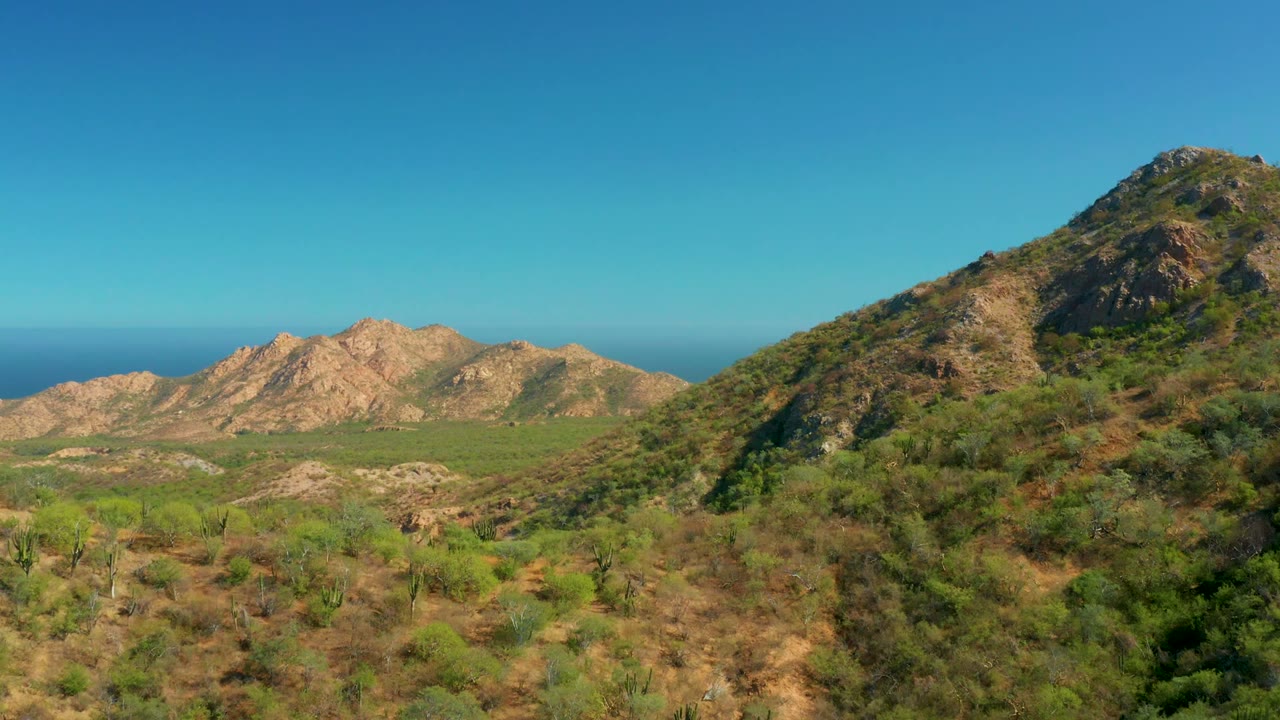 El Salado Viejo Los Cabos landscape with the right hillside occupying more of the frame, green desert vegetation below, rugged tan hills at left, distant ocean, and no visible people.