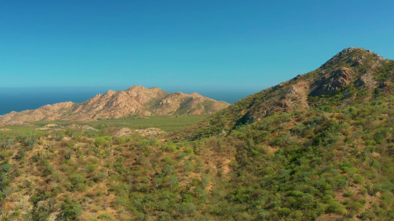 Opening frame of El Salado Viejo Los Cabos with scrub-covered rocky hills, tan ridges, a distant blue ocean horizon, and no visible people.