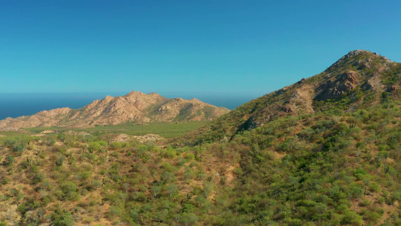 Aerial view across dry green hills toward tan mountains and ocean