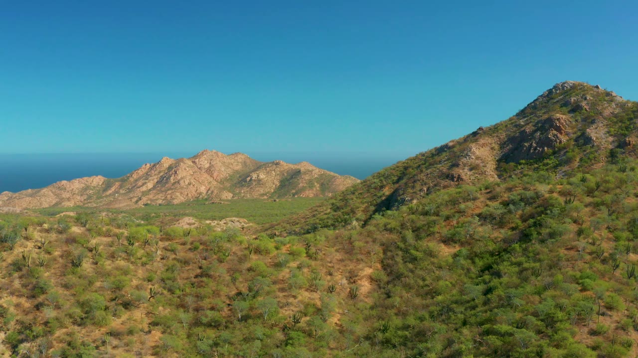 At 0:02, a taller scrub-covered slope enters strongly on the right of the El Salado Viejo Los Cabos frame, with lower tan ridges and ocean behind it, and no visible people.