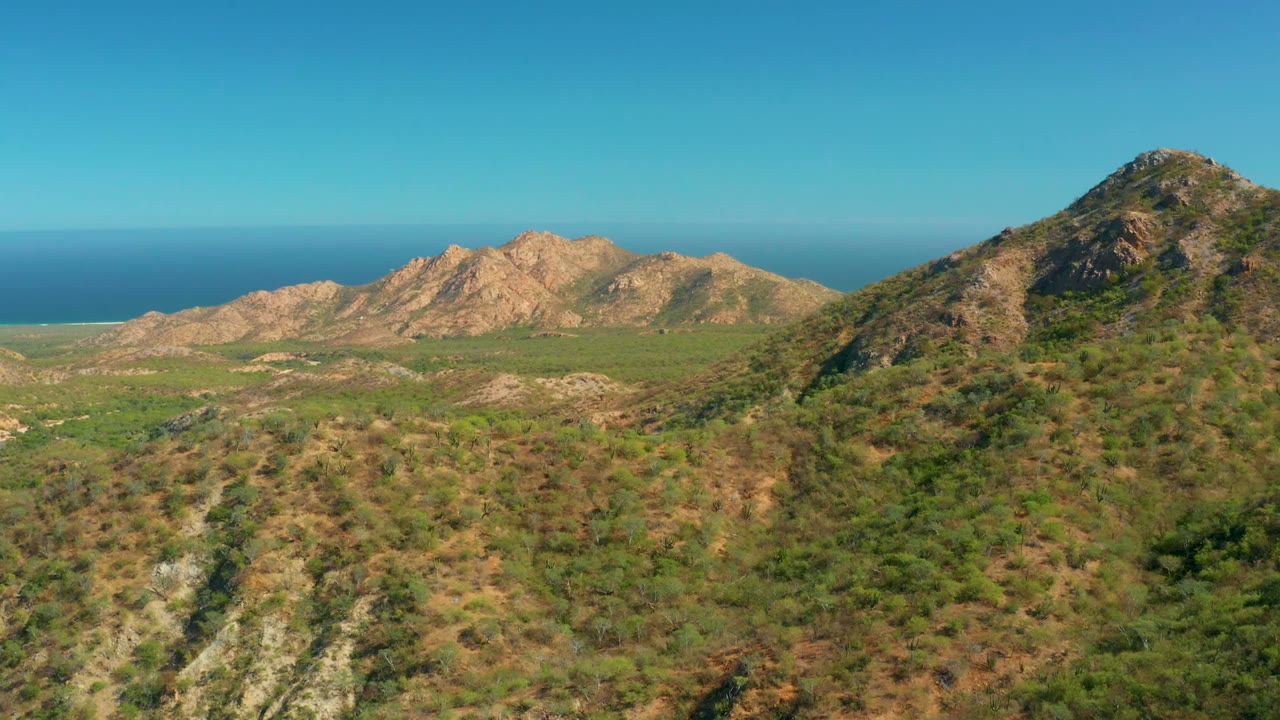 Aerial view over green-brown hills and tan rocky ridges near El Salado Viejo Los Cabos, with the blue ocean on the horizon and no visible people.