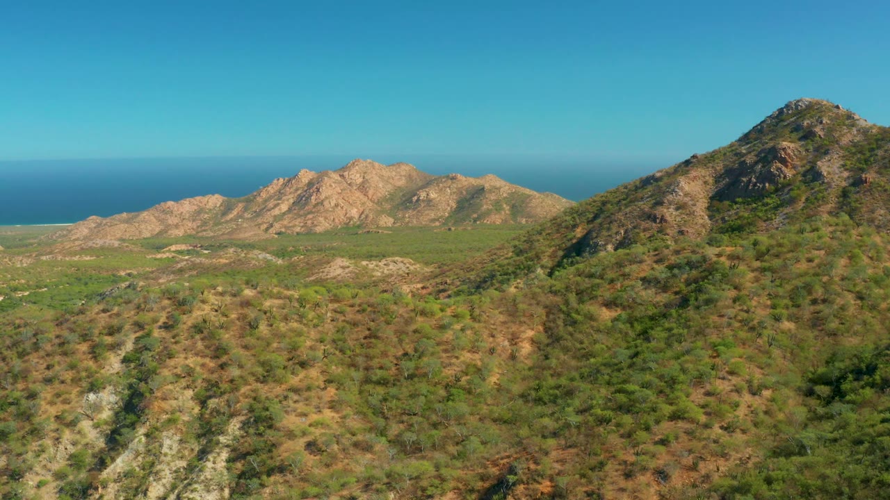 Rocky ridges, scrub-covered hills, and distant ocean under clear sky.
