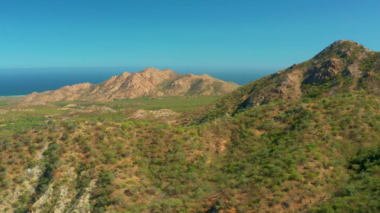 Aerial composition in El Salado Viejo Los Cabos with a nearer rocky green hillside at right, lower valley terrain at left, blue ocean in back, and no visible people.