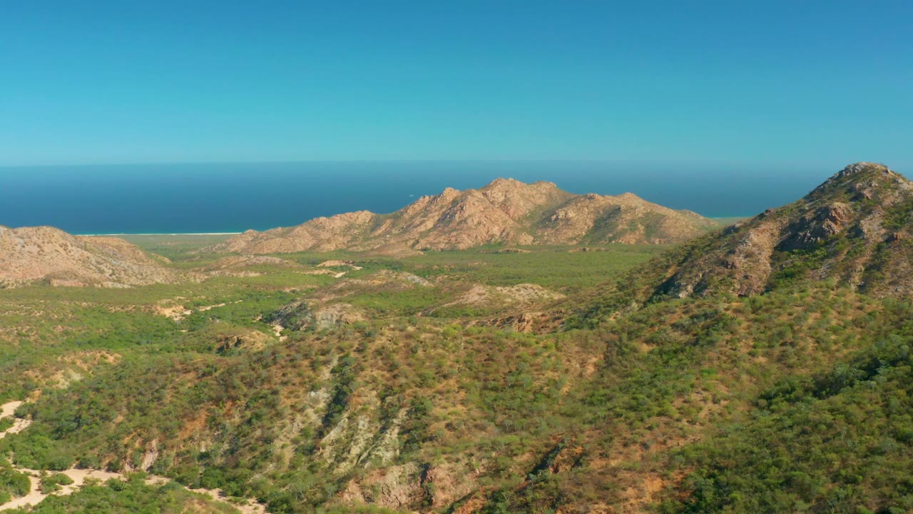 Opening frame of El Salado Viejo Los Cabos showing scrub-covered foreground hills, rocky tan ridges, a blue ocean horizon, clear sky, and no visible people.