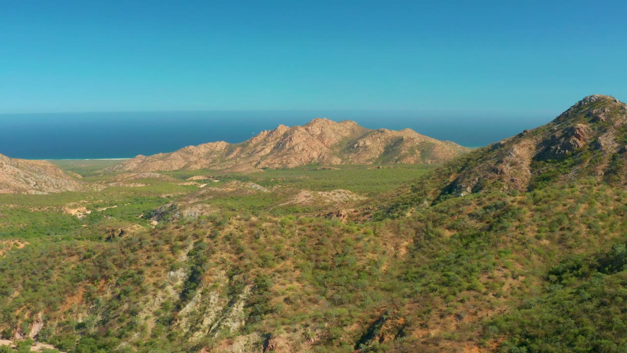 Opening aerial view over rocky green-brown hills in El Salado Viejo, Los Cabos, with a mountain ridge and blue ocean beyond, no visible people.