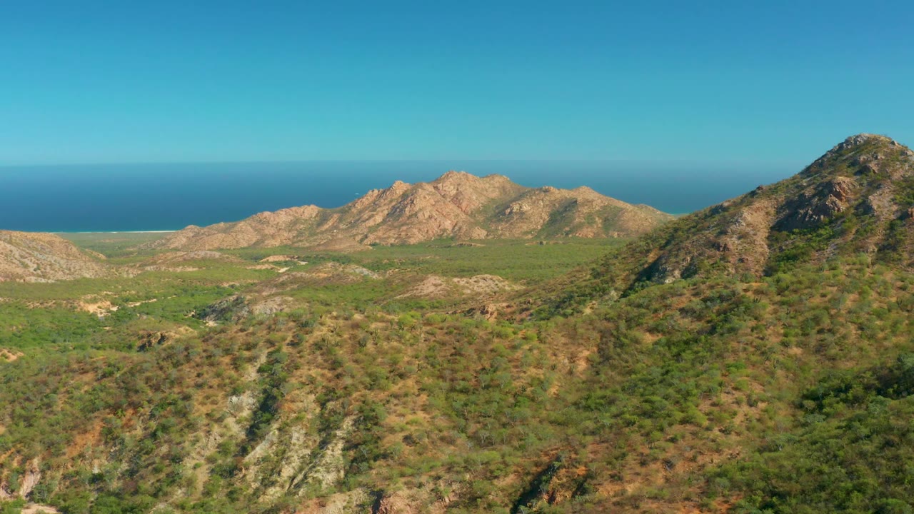 Aerial view of scrub-covered hills, distant mountains, and a blue ocean horizon.