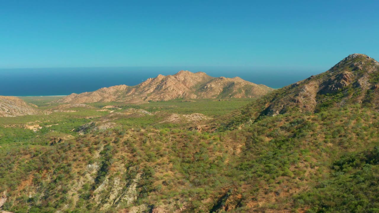Aerial frame over El Salado Viejo, Los Cabos, with a steep green-brown hill on the right, lower valley terrain across the center, and a blue ocean horizon, no visible people.