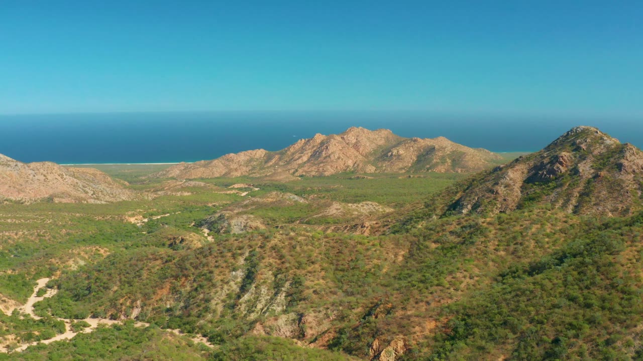 Opening frame over El Salado Viejo Los Cabos showing green hills, tan rocky ridges, blue ocean horizon, clear sky, and no visible people.