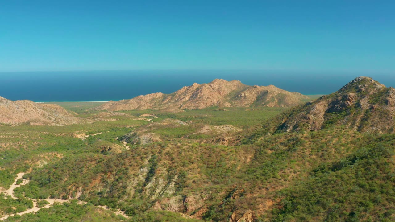 Third frame from the El Salado Viejo Los Cabos clip showing a darker foreground hill, pale dirt paths, tan ridges, blue sea in the distance, and no visible people.