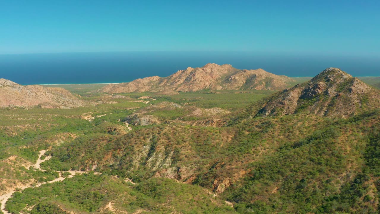 Aerial frame over El Salado Viejo Los Cabos showing green valley slopes, rocky ridges, a small white object in the sky area, blue ocean on the horizon, and no visible people.